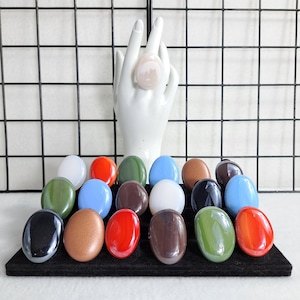 May include: A collection of polished, oval-shaped stones in various colors, including red, green, blue, brown, and black. A white ceramic hand holds a light pink stone. The stones are displayed on a black surface against a grid background.
