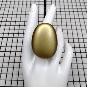 May include: A gold-colored ring with a smooth, oval-shaped design. The ring is displayed on a white mannequin hand.
