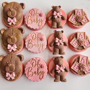 May include: An assortment of decorated cookies with a baby shower theme. The cookies feature teddy bear faces, teddy bear figures with pink bows, the words "Oh Baby" in gold script, and alphabet blocks with a rose accent. The cookies are pink, brown, and gold.