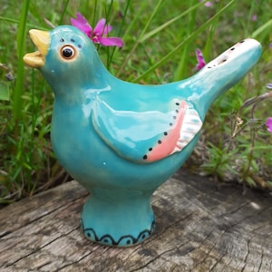 Op de afbeelding: Een turquoise keramieken vogelbeeldje met een open bek en een gele snavel. De vogel heeft een roze en wit vleugelontwerp met zwarte stippen. De basis van de vogel heeft een zwart golvend patroon. De vogel staat tegen een natuurlijke achtergrond van hout en groen.