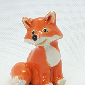 Peut inclure: Une figurine en céramique d'un renard assis avec sa queue enroulée derrière lui. Le renard est orange avec des accents blancs sur sa poitrine et sa queue. Le renard a une expression amicale avec des yeux noirs et un nez noir.