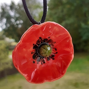Op de afbeelding: Een rode keramische klaproos hanger ketting met een zwart koord. De klaproos heeft een zwart midden met een groen midden en zwarte stippen.