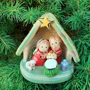 Op de afbeelding: Een keramische kerststalfiguur met Maria, Jozef en het kindje Jezus onder een groen dak met een gouden ster. Een lam en een blauwe lantaarn zijn ook inbegrepen. Het beeldje staat tegen een achtergrond van groene dennennaalden.