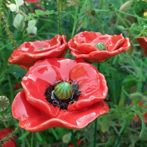 Op de afbeelding: Close-up van keramische rode klaprozen met groene centra en zwarte accenten. De bloemen staan tegen een achtergrond van groen gebladerte en andere bloemen. De klaprozen hebben een glanzende afwerking en zijn gerangschikt in een tuin.