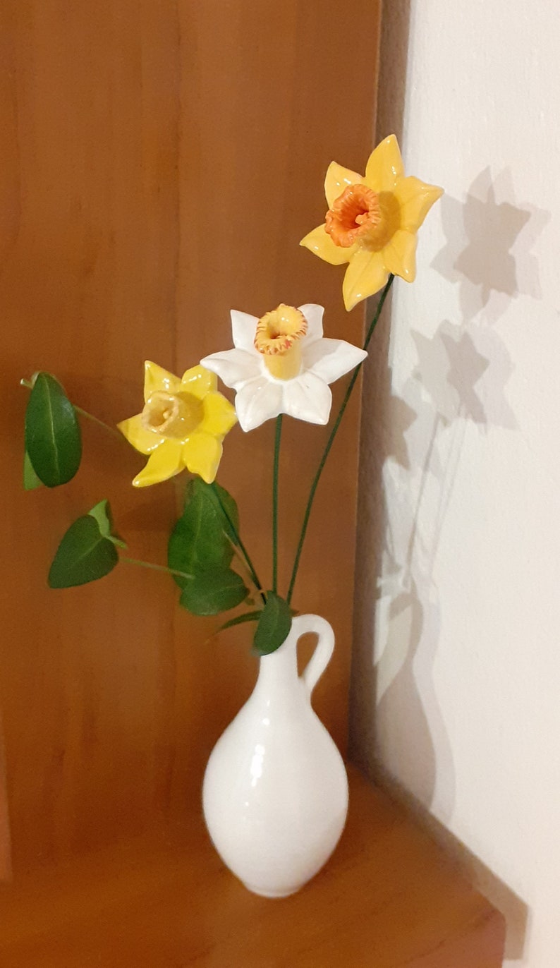 May include: A white ceramic vase with a narrow neck and a handle holds a bouquet of three yellow and white daffodils with green leaves.