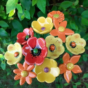 Op de afbeelding: Een verzameling kleurrijke keramische bloemensculpturen in rood, geel en oranje. De bloemen hebben gedetailleerde bloemblaadjes en zijn geplaatst tegen een achtergrond van groen gebladerte. Sommige bloemen hebben een lieveheersbeestjesontwerp in het midden.