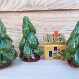 Puede incluir: Tres peque&ntilde;os pinos de cer&aacute;mica verde y una peque&ntilde;a casa de cer&aacute;mica amarilla con techo verde y chimenea naranja. La casa tiene ventanas azules y flores rojas.