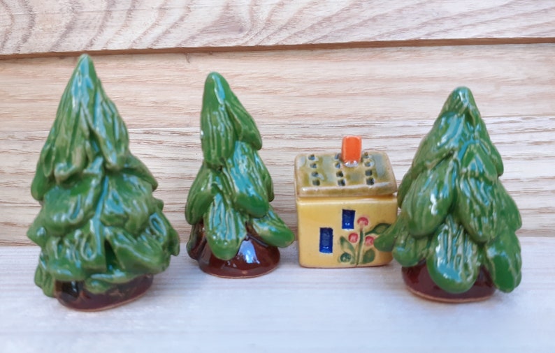 Puede incluir: Tres peque&ntilde;os pinos de cer&aacute;mica verde y una peque&ntilde;a casa de cer&aacute;mica amarilla con techo verde y chimenea naranja. La casa tiene ventanas azules y flores rojas.