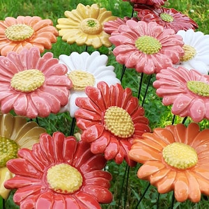 Op de afbeelding: Een verzameling kleurrijke keramische madeliefjes bloemtuinstokken. De bloemen zijn in tinten oranje, roze, rood, geel en wit, met gele centra. De stokken staan in groen gras.