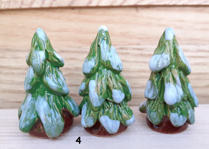 Puede incluir: Tres peque&ntilde;os &aacute;rboles de Navidad de cer&aacute;mica pintados en verde y blanco, cada uno con una base marr&oacute;n. Los &aacute;rboles est&aacute;n decorados con acentos blancos que parecen nieve.