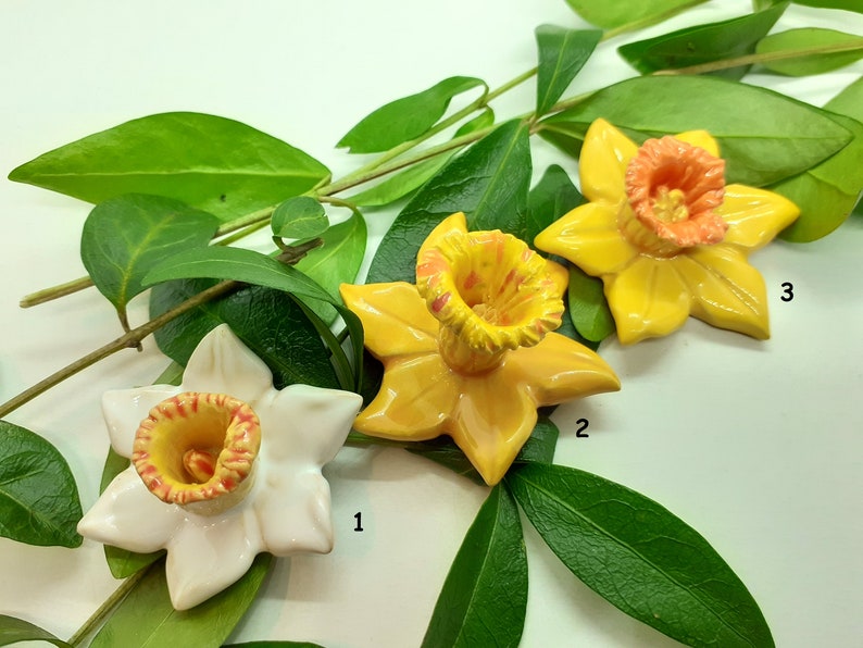 May include: Three ceramic daffodil flower sculptures in yellow and orange. The flowers are arranged on a green leafy branch. The flowers are numbered 1, 2, and 3.