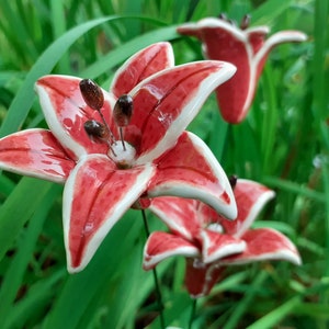Op de afbeelding: Drie rode en witte keramische leliebloemen met groene stelen. De lelies zijn in een cluster gerangschikt en zijn geplaatst tegen een achtergrond van groen gras.