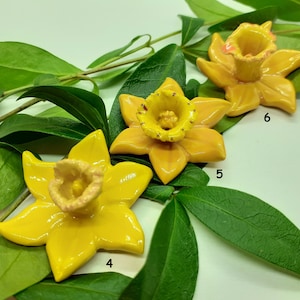 May include: Three yellow ceramic daffodil flower sculptures on a green leafy background. The flowers are arranged in a row, with the largest flower in the center. The flowers have a yellow glaze and a pink center.
