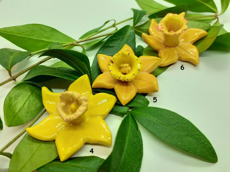 May include: Three yellow ceramic daffodil flower sculptures on a green leafy background. The flowers are arranged in a row, with the largest flower in the center. The flowers have a yellow glaze and a pink center.