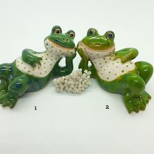 Op de afbeelding: Twee keramische kikkerbeeldjes met groene en witte stippenpatronen. De kikkers zitten met gekruiste armen en hebben een ondeugende uitdrukking op hun gezicht.