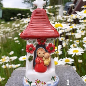 Op de afbeelding: Een keramieken religieus beeldje dat een kapel met een rood dak en een wit kruis voorstelt. De kapel toont een afbeelding van Maria en het kindje Jezus, met rode bloemaccenten. De basis is versierd met bloemmotieven en blauwe accenten.