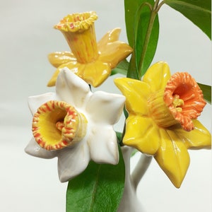 May include: Three ceramic daffodils in a white vase. The daffodils are yellow with orange centers. The vase is white and has a round shape.