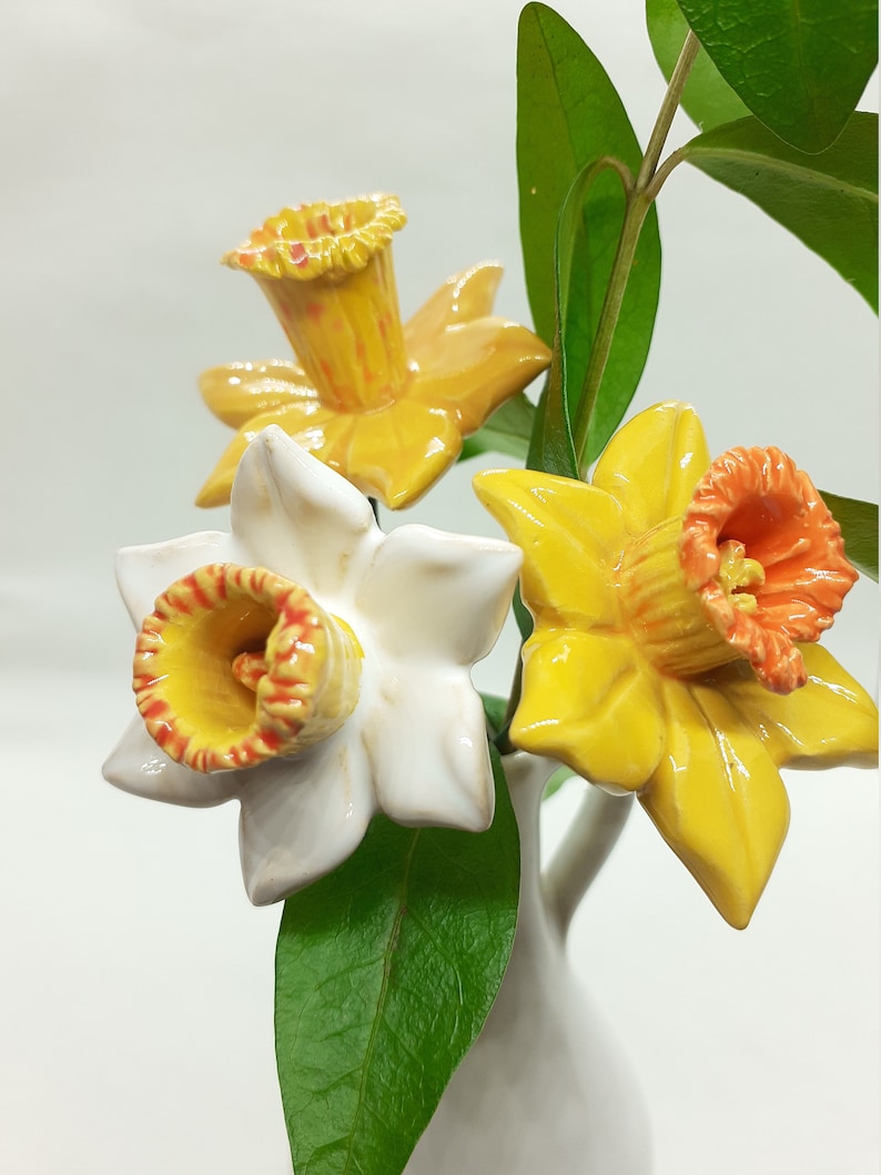 May include: Three ceramic daffodils in a white vase. The daffodils are yellow with orange centers. The vase is white and has a round shape.
