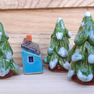 Puede incluir: Un conjunto de cuatro adornos de &aacute;rbol de Navidad de cer&aacute;mica, cada uno con un esmalte verde y blanco, y una peque&ntilde;a casa de cer&aacute;mica azul con un techo marr&oacute;n y una chimenea naranja.