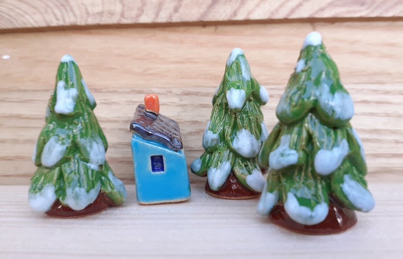Puede incluir: Un conjunto de cuatro adornos de &aacute;rbol de Navidad de cer&aacute;mica, cada uno con un esmalte verde y blanco, y una peque&ntilde;a casa de cer&aacute;mica azul con un techo marr&oacute;n y una chimenea naranja.