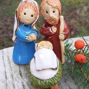 Op de afbeelding: Een kerststal van keramiek met een Maria in een blauwe robe, een Jozef in een rode robe en een baby Jezus in een kribbe. De figuren zijn handgeschilderd met gouden accenten en hebben een rustieke, vintage uitstraling.