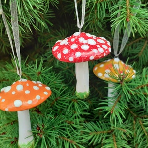 Op de afbeelding: Drie keramische paddenstoelversieringen die aan een kerstboom hangen. De paddenstoelen zijn rood met witte stippen, oranje met witte stippen en geel met witte stippen.