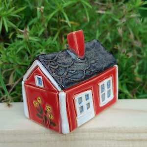 Op de afbeelding: Een miniatuur rood keramisch huis met een donkergrijs getextureerd dak en een rode schoorsteen. Het huis heeft witte randen rond de ramen en deuren, met een bloemenontwerp aan de zijkant. De ramen hebben blauwe ruiten.