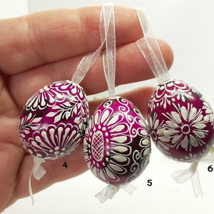 Op de afbeelding: Drie roze paaseieren met witte bloemmotieven en zwarte accenten. De eieren hangen aan witte linten en zijn genummerd 4, 5 en 6.