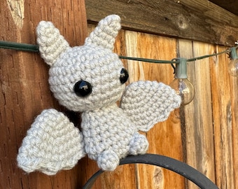 May include: A gray crocheted bat with black eyes hanging on a wooden fence with string lights.