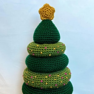 May include: A green crocheted Christmas tree toy with a brown base and a gold star on top. The tree is decorated with pink and yellow beads.