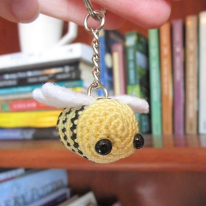 Puede incluir: Un llavero de abeja de crochet con alas blancas, rayas negras y ojos negros. La abeja está unida a una cadena plateada y un llavero.