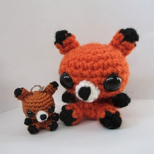 May include: Two crocheted fox plushies, one large and one small. The larger fox is orange with black ears and paws, and white accents around the nose and mouth. The smaller fox is a keychain with the same design.