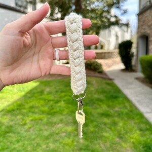 May include: A white crocheted keychain with a silver key attached. The keychain is held in a hand against a blurred background of green grass and a building.
