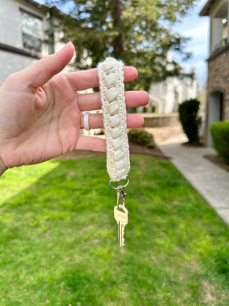 May include: A white crocheted keychain with a silver key attached. The keychain is held in a hand against a blurred background of green grass and a building.