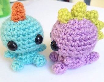 May include: Two crocheted amigurumi toys, one blue with an orange horn and the other purple with a green crest. Both have black eyes.