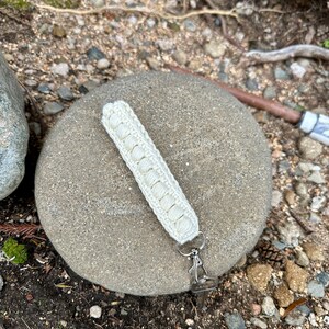 May include: A white crocheted keychain with a silver metal clasp. The keychain is lying on a gray rock.