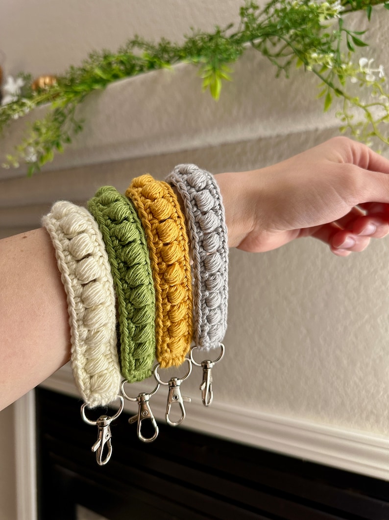 May include: Four crocheted keychains in different colors: cream, green, yellow, and gray. Each keychain has a silver metal clasp.
