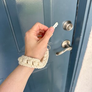 May include: A hand holding a key with a white crocheted keychain attached to it. The keychain is made of a series of small, round, white crocheted balls.
