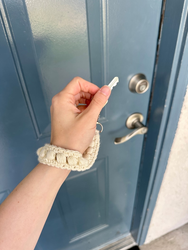 May include: A hand holding a key with a white crocheted keychain attached to it. The keychain is made of a series of small, round, white crocheted balls.