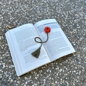 May include: A green and brown crocheted bookmark with a tassel and a red flower on top. The bookmark is placed on an open book with the text "Create Meaningful Experiences" visible.