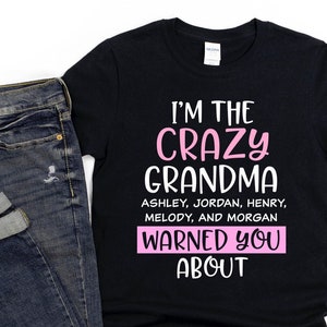 May include: Black t-shirt with the text "I'M THE CRAZY GRANDMA" in pink and white. The names Ashley, Jordan, Henry, Melody, and Morgan are listed below. Rolled-up blue jeans are next to the shirt.