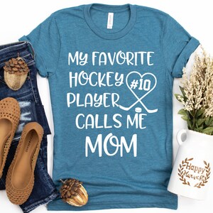 May include: A teal blue t-shirt with the text "My favorite hockey player #10 calls me mom" in white. The text is surrounded by a heart and hockey sticks.