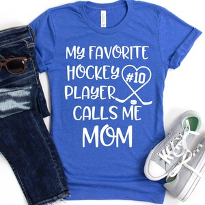 May include: A royal blue t-shirt with white text that reads "My favorite hockey player #10 calls me mom". The text is accompanied by a white heart with the number 10 inside, two hockey sticks crossed over each other, and a hockey puck.