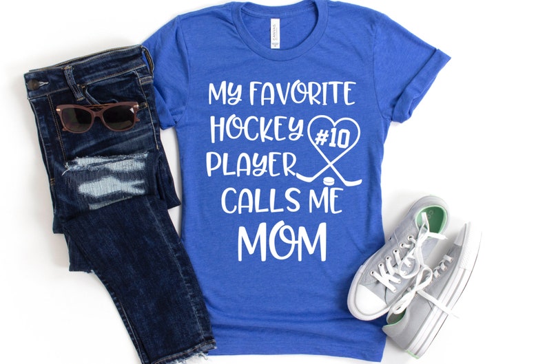 May include: A royal blue t-shirt with white text that reads "My favorite hockey player #10 calls me mom". The text is accompanied by a white heart with the number 10 inside, two hockey sticks crossed over each other, and a hockey puck.