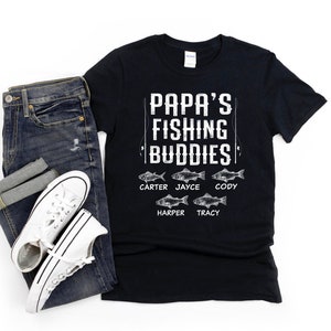 Puede incluir: Camiseta negra con texto blanco que dice "Papa's Fishing Buddies" con un gr&aacute;fico de ca&ntilde;a de pescar y cinco peces con nombres debajo de cada pez: Carter, Jayce, Cody, Harper y Tracy.