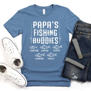 Puede incluir: Una camiseta azul con el texto "Papa's Fishing Buddies" y los nombres Carter, Jayce, Cody, Harper y Tracy impresos debajo. Cada nombre est&aacute; emparejado con una ilustraci&oacute;n de un pez.