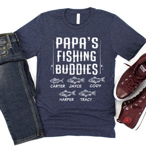 Puede incluir: Una camiseta azul marino con el texto "Papa's Fishing Buddies" y cinco peces con nombres debajo: Carter, Jayce, Cody, Harper y Tracy.