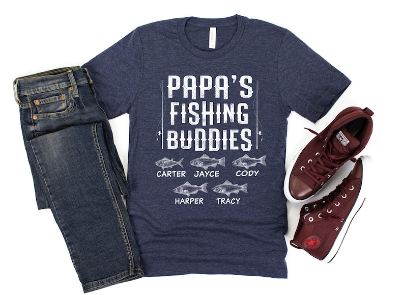 Puede incluir: Una camiseta azul marino con el texto "Papa's Fishing Buddies" y cinco peces con nombres debajo: Carter, Jayce, Cody, Harper y Tracy.