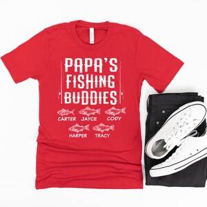 Puede incluir: Camiseta roja con texto blanco que dice "Papa's Fishing Buddies" con cinco peces y nombres debajo de cada pez: Carter, Jayce, Cody, Harper y Tracy.