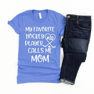 May include: A blue t-shirt with white text that reads "My favorite hockey player #10 calls me mom". The text is in a heart shape with hockey sticks and a puck.